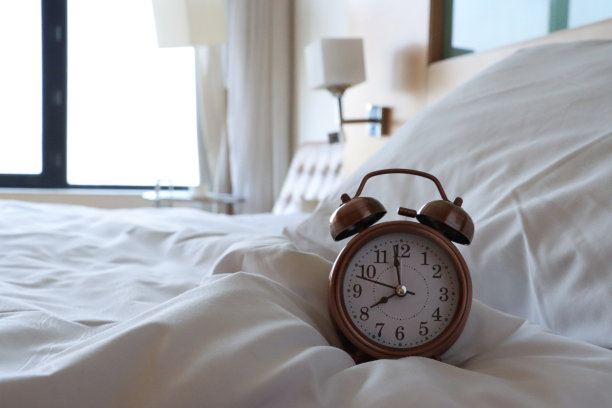 青铜金属案件的特写图像，复古闹钟，双钟，模拟显示时钟与白色表盘坐在白色羽绒被床罩，黑色数字，小时和分针，枕头靠在木头和玻璃床头板，重点在前景图片下载