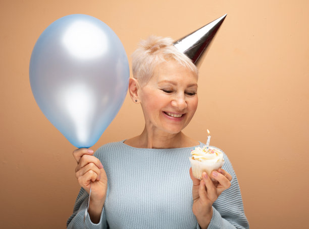 美丽的老年女性手里拿着带蜡烛的纸杯蛋糕和蓝色气球。图片下载