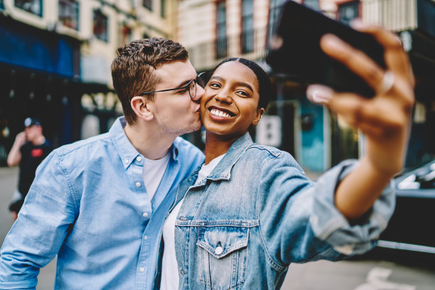 白人潮男亲吻快乐的黑皮肤女友，同时摆姿势和自拍，在一起旅行中，通过手机前置摄像头拍摄真实多样的婚姻图片下载