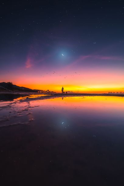 人站在岸边，月亮和太阳的倒影在平静的水面上图片下载