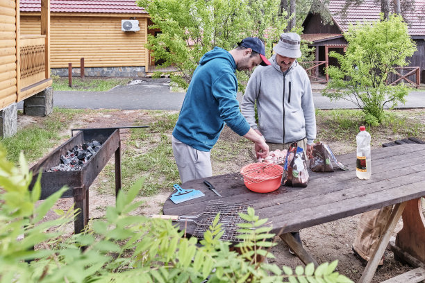 亚洲父亲和成年的儿子在后院烧烤时把生鸡肉片放在烤架上。图片下载