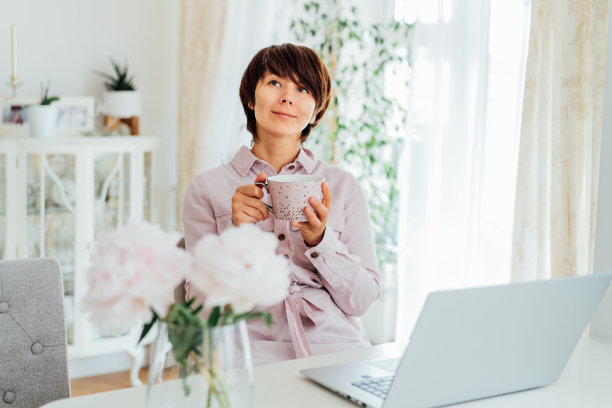 做梦的女人在家里的笔记本电脑上喝一杯放松的工作。舒适的工作空间，粉红色的牡丹花在桌子上，室内光线充足。对所做的工作感到满意。需要时间来恢复。图片下载