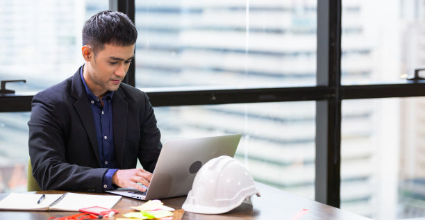 亚洲男性工程师或建筑师坐在办公室的办公桌上用笔记本电脑工作。严肃的工作。把安全帽放在桌上。图片下载