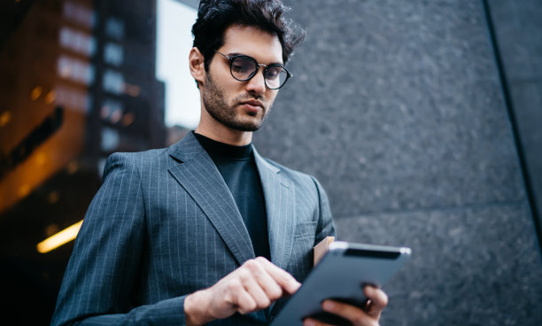 成功的男性企业家穿着优雅的服装，在连接4G网络的现代触摸板设备上下载带有商业新闻的文件，站在办公楼附近，烘焙浏览图片下载