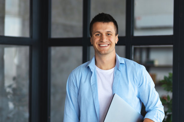 成功的年轻商人或企业家站在现代办公室的肖像。快乐的程序员用笔记本电脑看着镜头，友好地微笑着图片下载