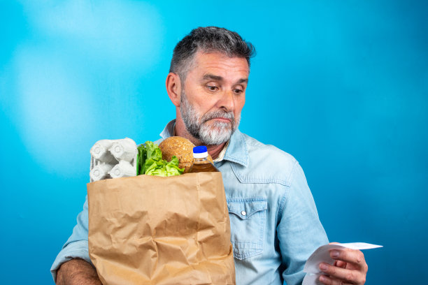 成年男子购物后震惊地看着商店收据，手里拿着一个装着健康食品的纸袋。真实的人的表情。通货膨胀的概念。拿着一纸袋杂货的男人看起来很惊讶。图片下载