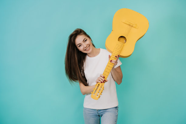 美丽的金发年轻女子疯狂的表情在白色t恤尖叫上升黄色吉他想打破它，在蓝绿色的工作室背景。漂亮的意大利女孩拿着吉他。模型图片下载