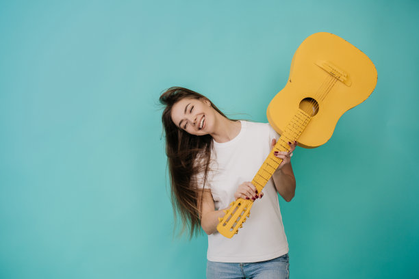 美丽的金发年轻女子与漂亮的微笑在白色t恤笑闭眼睛上升黄色吉他想打破它，在蓝绿色的工作室背景。漂亮的意大利女孩拿着吉他。模型图片下载