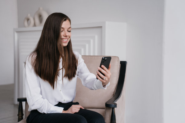 感激的意大利黑发年轻女子视频通话使用电话坐在椅子上，笑容灿烂。学生女孩通过互联网与朋友打招呼。成功的企业家。图片下载