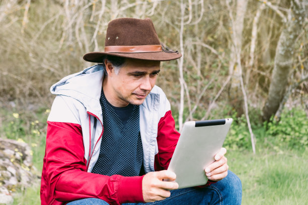 一位戴着帽子的企业家，坐在一块石头上，一边放松一边看着他的平板电脑。工作、享受、放松、互联网、技术和连接的概念。图片下载