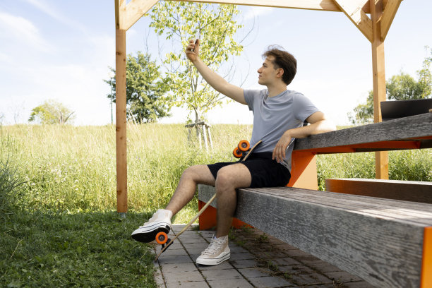 年轻人坐在公园的长椅上，拿着滑板和智能手机图片下载