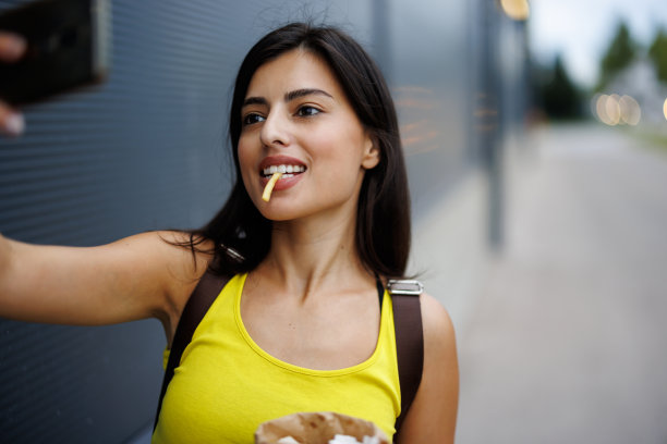 美丽的年轻女子一边走在城市里，一边吃着薯条，用手机自拍图片下载
