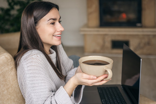 一个年轻的女人拿着一大杯咖啡，笔记本电脑放在家里的沙发上。图片下载