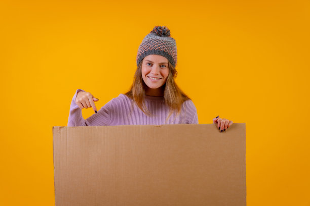 一个戴着羊毛帽的女人指着黄色背景上的纸板招牌图片下载