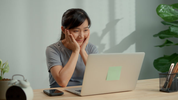 亚洲妇女开心地微笑着在家里办公室用笔记本电脑看电影。图片下载