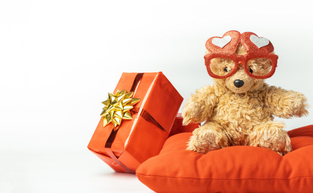 小毛绒泰迪熊戴着红色心形眼镜，拿着红色气球。情人节快乐。礼物用红色包装纸包装，白色背景上点缀着金色蝴蝶结。图片下载