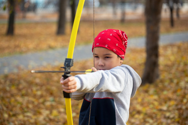 亚洲7岁男孩在一个红色的大手帕瞄准与弓户外公园在秋天，近距离，选择性聚焦。图片下载