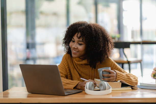 非裔美国女孩用笔记本电脑坐在书桌前写作。努力工作。女子在网上工作时戴着耳机。学生兴奋地在咖啡馆在线学习语言。图片下载