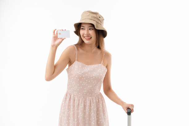 微笑的年轻亚洲女性旅行者，穿着粉红色的连衣裙和帽子，拿着信用卡和白色背景上孤立的行李箱。图片下载