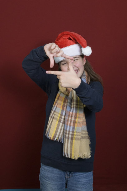 一个戴着圣诞老人帽子的女孩在做一个手势——这是照片胶片的象征。暗红色背景。垂直图片下载