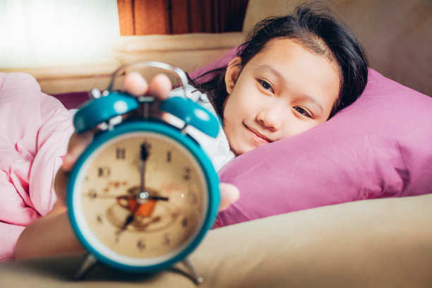 快乐的醒来，一个美丽的女孩躺在床上，闹钟停了。清晨醒来。学生们很早就起床去上学。图片下载