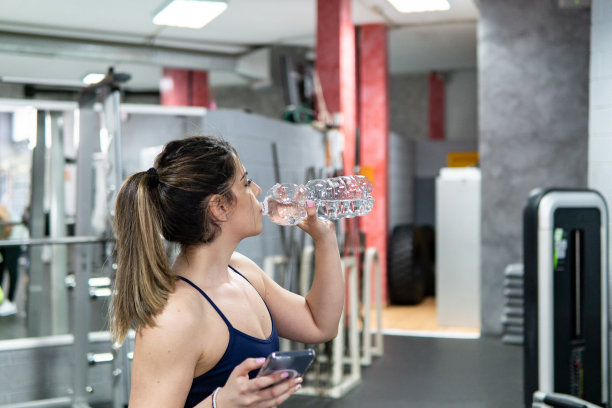 年轻的白人女子拿着智能手机在健身房休息，锻炼后喝水图片下载