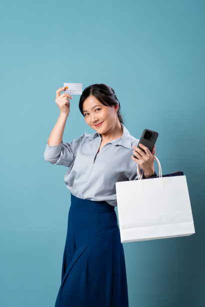 亚洲女性快乐的微笑着拿着智能手机，信用卡和购物袋隔离在蓝色背景。图片下载