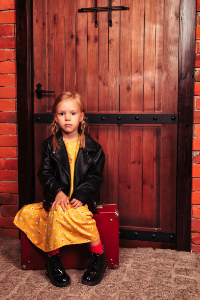 穿着皮夹克的学龄前女孩坐在木门背景的乡村行李箱上，看着相机。小模特脸上带着消极情绪，沉思。时尚风格概念。复制文本空格图片下载