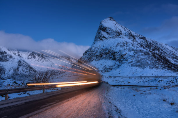 冬天的夜晚，路上的雪山和模糊的汽车前灯。罗弗敦群岛，挪威。美丽的风景与岩石在雪，云，蓝天和道路在黄昏。光线轨迹。自然图片下载