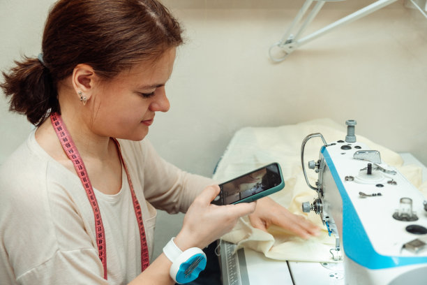 一名白人女裁缝在家庭作坊的缝纫机上缝纫，用智能手机录下广播。小生意。博客的概念图片下载