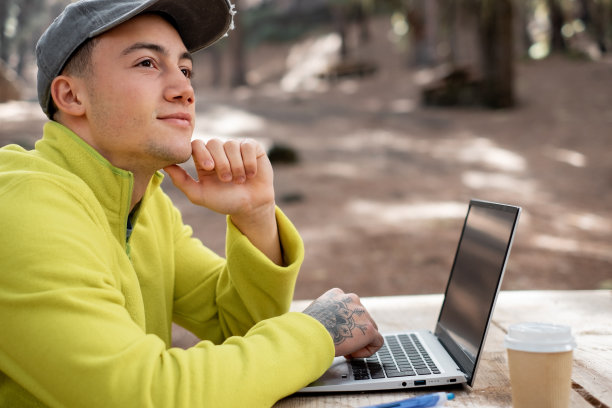 在新鲜空气中做生意。沉思美丽的年轻男子在笔记本电脑上工作，坐在森林里的户外木桌旁。自然智能工作的概念图片下载