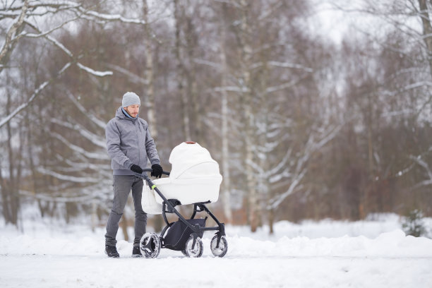 微笑的年轻成年父亲推着白色的婴儿车，在寒冷的冬天走在公园积雪覆盖的人行道上。陪伴新生儿，呼吸新鲜空气。享受宁静的散步。图片下载