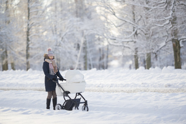 在寒冷的冬天，年轻的成年母亲推着白色的婴儿车走在公园积雪覆盖的人行道上。陪伴新生儿，呼吸新鲜空气。享受宁静的散步。图片下载