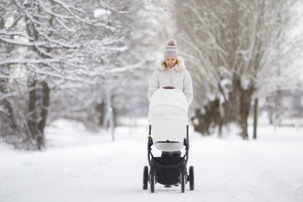 在寒冷的冬天，年轻的成年母亲推着白色的婴儿车走在公园积雪覆盖的人行道上。陪伴新生儿，呼吸新鲜空气。享受宁静的散步。图片下载