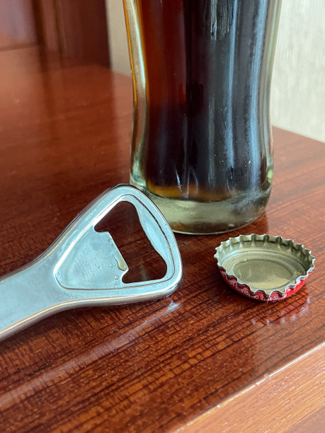 餐厅场景图片，桌子上放着玻璃瓶可乐，开瓶器躺在旁边，取下金属瓶盖，聚焦前景图片下载