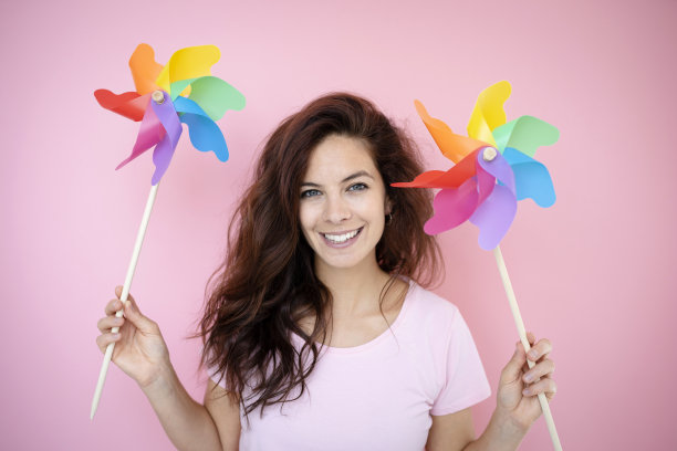 漂亮的年轻女子拿着两个五颜六色的彩虹风车，站在粉红色的背景前面图片下载