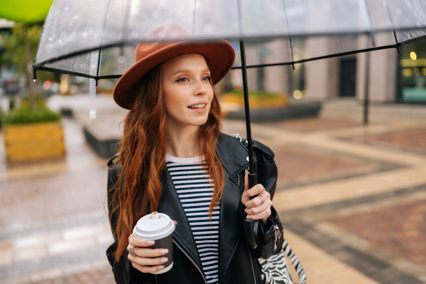 在寒冷的秋雨中，快乐的年轻女子戴着时尚帽，手里拿着一杯热咖啡，站在透明的雨伞下。图片下载