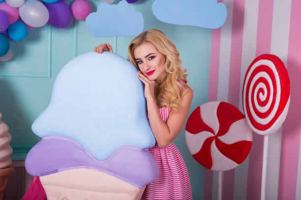 画像的年轻女子在粉红色的裙子拿着大冰淇淋和摆造型装饰的背景。可爱的糖果女孩被玩具糖果包围。图片下载