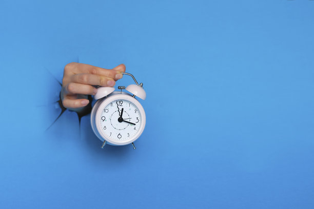 女性手拿粉色闹钟穿过蓝色背景的纸洞。图片下载