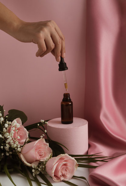 讲台上的棕色玻璃滴管瓶，粉红色的玫瑰花和女人的手。帕特尔粉丝质窗帘背景。护肤产品，天然化妆品。美丽的概念。图片下载