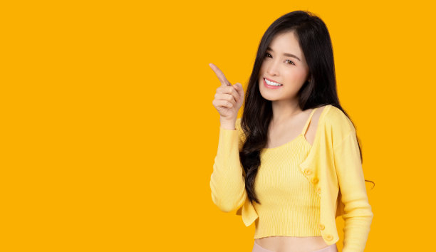 美丽年轻的亚洲女子指向复制空间，看着相机微笑的脸和幸福漂亮的女孩表现得像一个满意的产品，用于广告与孤立的黄色背景图片下载