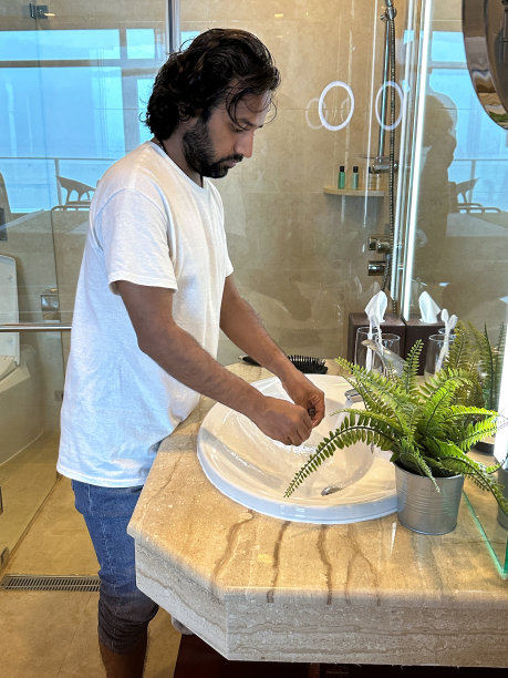 印度男人站在豪华浴室的水槽反射的镜像，穿着白色毛巾长袍和毛巾包裹着头，在自来水下洗手，棕色马赛克瓷砖墙背景，重点在前景图片下载