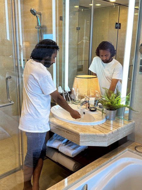 印度男人站在豪华浴室的水槽里，在自来水下洗手，在大理石浴室柜台上的金属锅里种植人造蕨类植物，流动的水龙头，后视图，关注前景图片下载