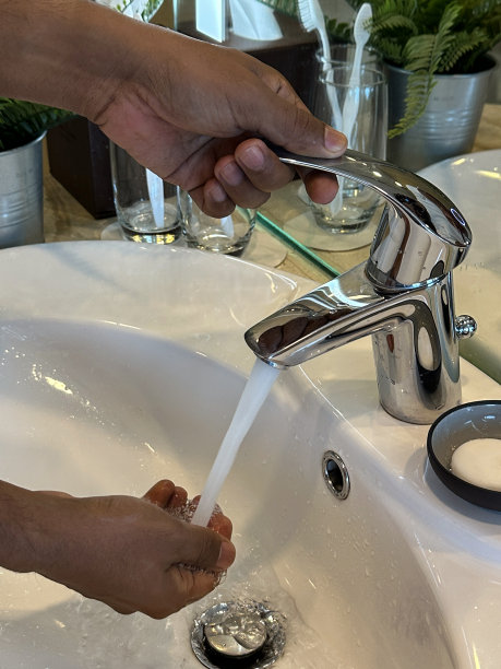 特写图像，一个无法辨认的人站在豪华浴室的水槽下，在自来水下洗手，大理石浴室柜台上的金属花盆里的人造蕨类植物，流动的水龙头，侧面视图，关注前景图片下载