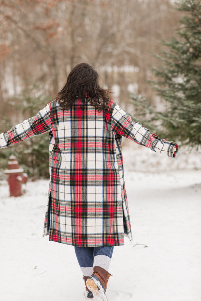 2022年12月，俄亥俄州杰克逊，一名31岁的棕色女子在新鲜的白色蓬松的雪中站在一棵真正的圣诞树旁，她穿着鸭皮靴和圣诞格子格纹大衣，手臂伸出图片下载