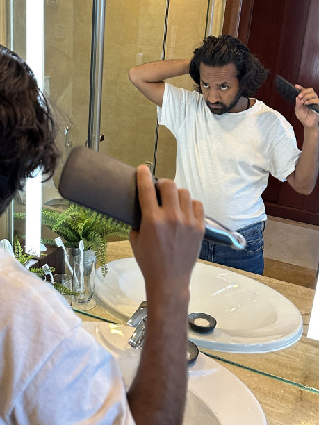 镜像反射的印度男人造型凌乱的头发与梳子在浴室，头发护理，人造蕨类植物在金属锅在大理石浴室柜台，牙刷在玻璃，重点在前景图片下载