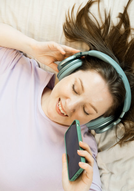 这名女子戴着耳机躺在床上，闭着眼睛听音乐，手里拿着智能手机图片下载