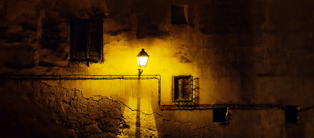 在一个老城区，一堵旧的、风化的、损坏的墙的正面，有窗户和金属栏杆，还有一盏昏暗的橙黄色灯光的古老路灯。令人毛骨悚然的中世纪壁纸背景的黑色明信片图片下载