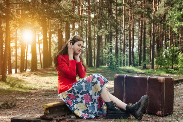 十几岁的女孩在时尚的复古形象与复古行李箱坐在铁路在夏天，听音乐耳机。森林铁路上的年轻时尚女士。音乐风格概念。复制文本空格图片下载