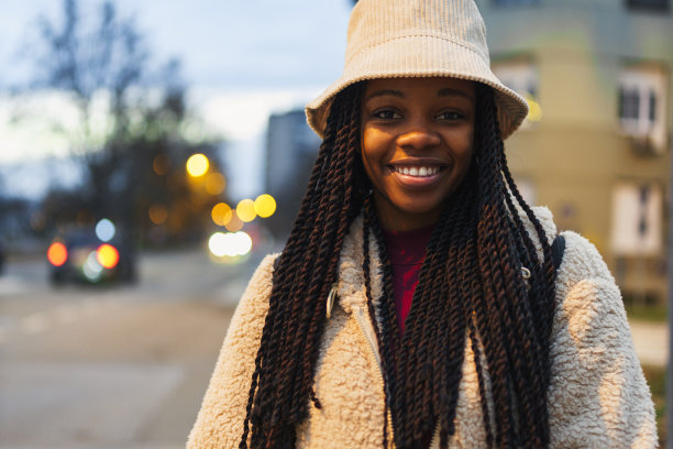 一个美丽的黑人女性对着镜头兴奋地微笑，她戴着水桶帽，背着背包，喝着咖啡，用她的手机拍照图片下载